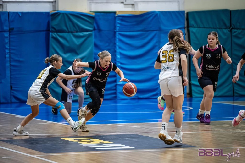 spēles moments. mūsu spēlētāja ar bumbu azartiski cīnās, divas pretinieces mēšina at nemt bumbu.