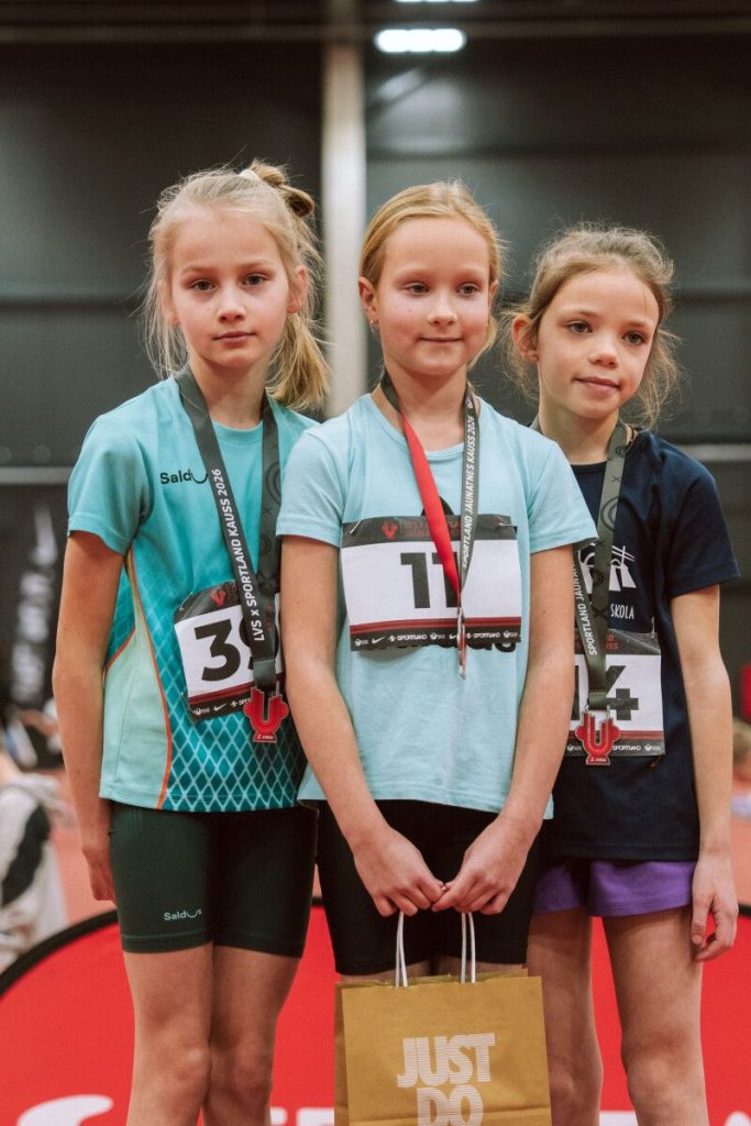 trīs meitenes uz pirmās pakāpes pjedestāla ar medaļām kaklā