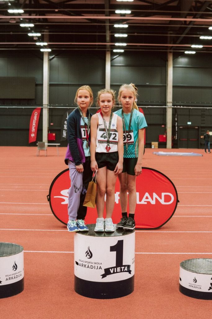 trīs meitenes uz pirmās pakāpes pjedestāla ar medaļām kaklā
