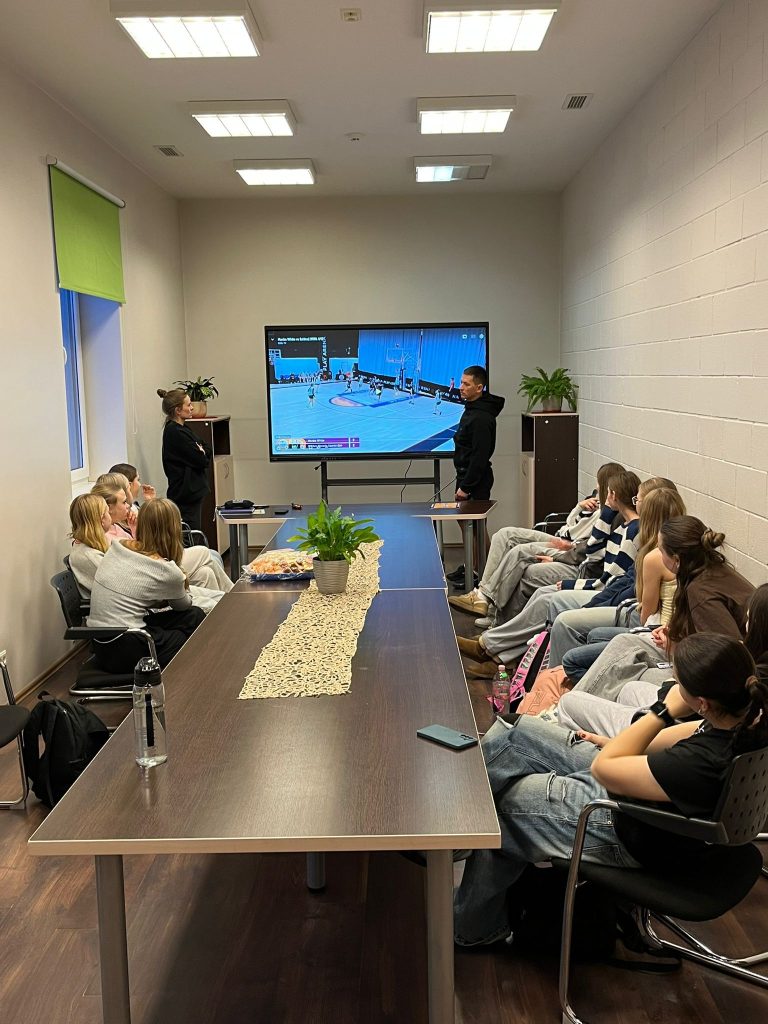 treneri stāv kājās pie ekrāna. Audzēkņi sasēdu