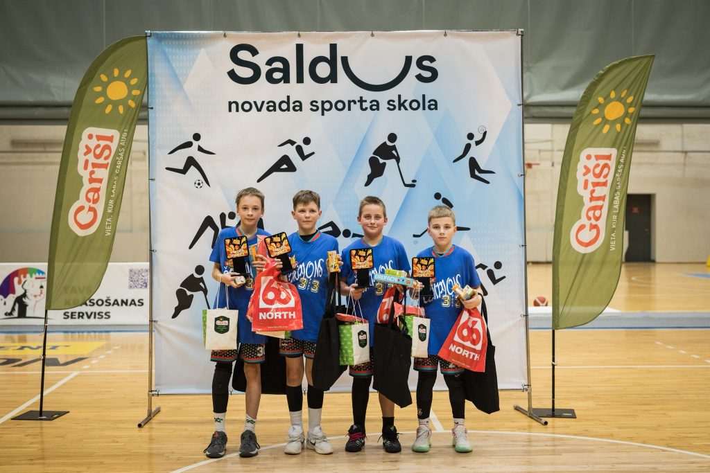 bērni pie sporta skolas banera, rokās dāvanu maisiņi, medaļas.