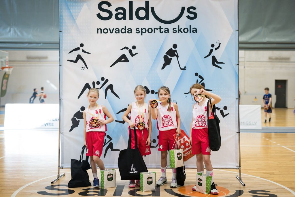 bērni pie sporta skolas banera, rokās dāvanu maisiņi, medaļas.