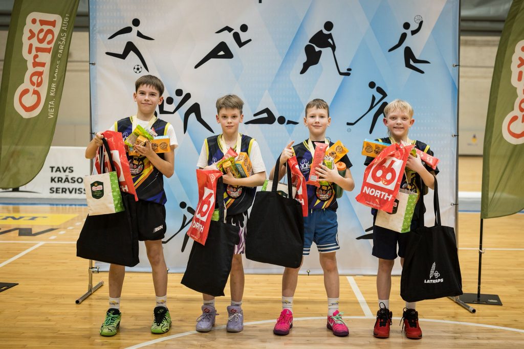 bērni pie sporta skolas banera, rokās dāvanu maisiņi, medaļas.