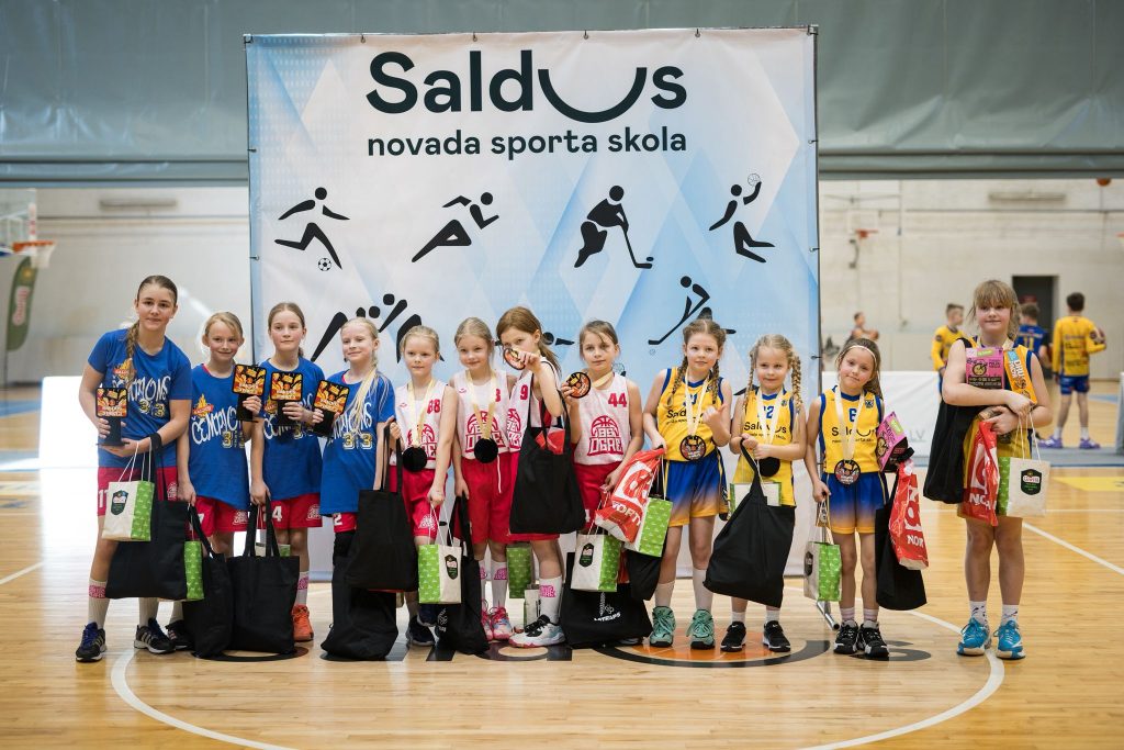 bērni pie sporta skolas banera, rokās dāvanu maisiņi, medaļas.