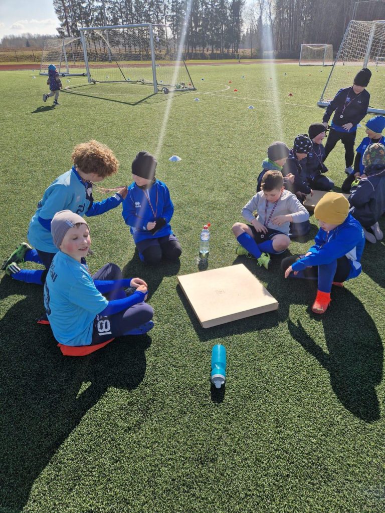 bērni apsēdušies zālītē - laukuma malā. Gatvaojas vērt vaļā picu kasti